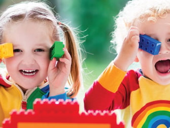 Two children in rainbow shirts hold colorful blocks to their eyes, smiling.