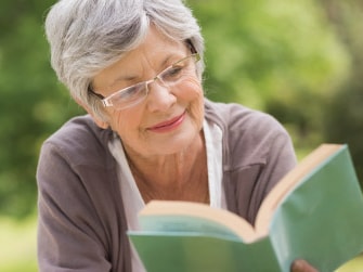 Elderly woman with glasses reading a green book outside amid lush greenery.