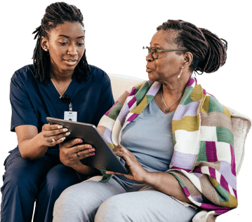 A woman in scrubs shows an older woman a tablet, both seated on a couch.