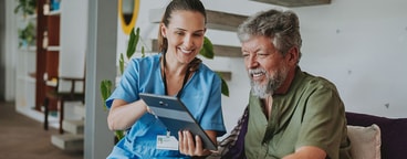 A healthcare worker and an older man smile while looking at a tablet in a bright room.