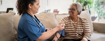 A medbridge healthcare worker checks an elderly woman's blood pressure on a couch.