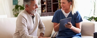 A Medbridge clinician in blue scrubs uses a tablet to consult with an older man.