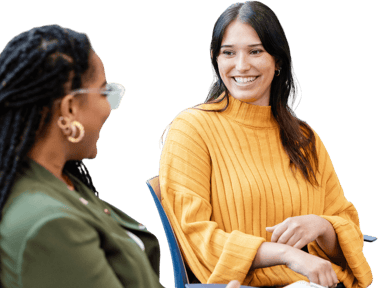 Two women sit and talk; one smiles in a yellow sweater, the other listens in green—possibly discussing digital healthcare or medbridge training.