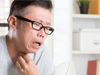 A person with glasses and a gray shirt touches their neck indoors, showing discomfort.
