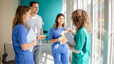 Four healthcare workers in scrubs talk by bright windows, holding clipboards with medbridge software visible.