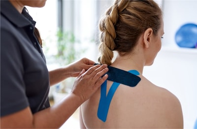 In a bright room, someone applies blue kinesiology tape to a woman's upper back and neck, blending traditional with modern care.