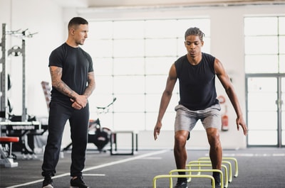 In a spacious gym, one man jumps hurdles under another's watchful eye, highlighting medbridge's commitment to fitness and wellness.