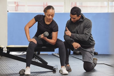 A gym scene shows a woman lifting a dumbbell with guidance from a man, highlighting hybrid care for wellness.