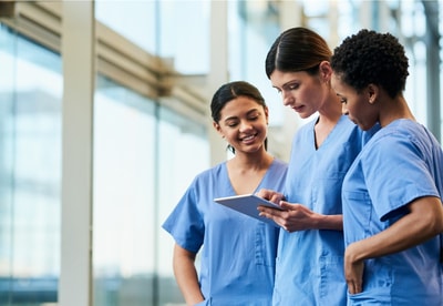 Three healthcare professionals in blue scrubs review Medbridge software on a tablet in a bright hallway.
