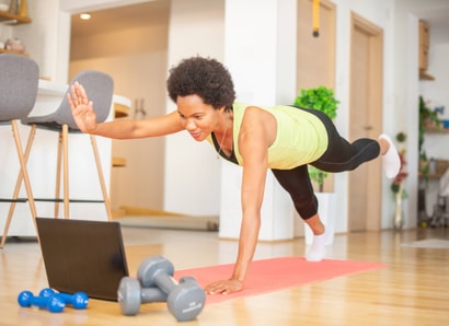 Woman in athletic wear does a plank on a yoga mat at home, following a medbridge workout on her laptop with dumbbells close by.