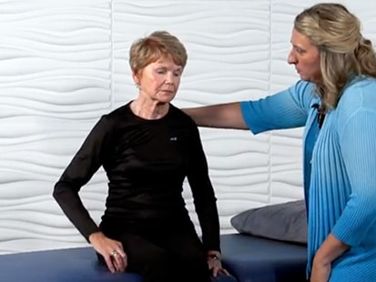 A Medbridge staff member in blue helps a person seated at a table against a wavy white wall.
