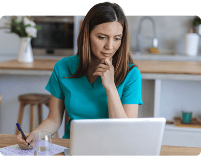 Woman in teal scrubs at kitchen table uses laptop for medbridge healthcare training, holding pen and paper.