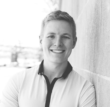 Person with short light hair leans against a wall, wearing a shirt with a dark collar in a black and white photo.
