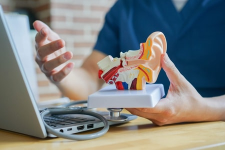 A person shows a model ear at a desk with a laptop and stethoscope, representing hybrid care and medbridge software.