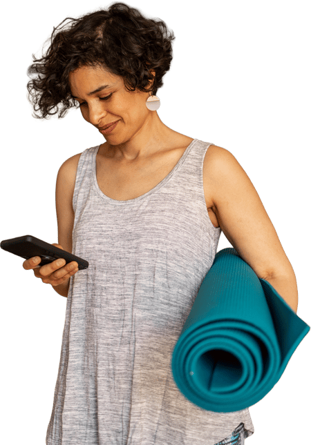 Woman in a gray tank top holds a yoga mat and checks her phone, possibly using medbridge.