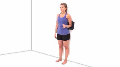 In a white room, a person in a blue tank top and black shorts stands barefoot with a cushion between their elbow and the wall, resembling healthcare training exercises from Medbridge.