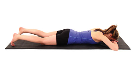 A person in a blue top and black shorts lies face down on a mat in a healthcare training pose.