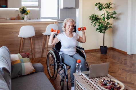 Older woman in a wheelchair exercises with dumbbells, following a medbridge workout on her laptop at home.