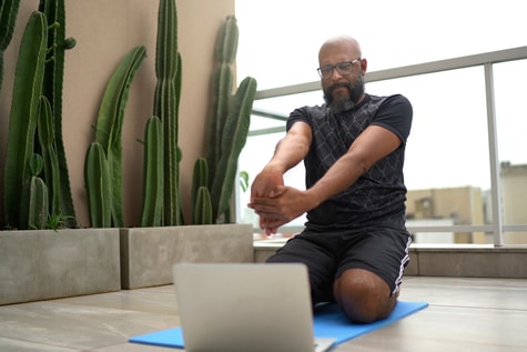 Man stretches on a yoga mat indoors, looking at a laptop, with cacti nearby—showing medbridge use for home wellness.