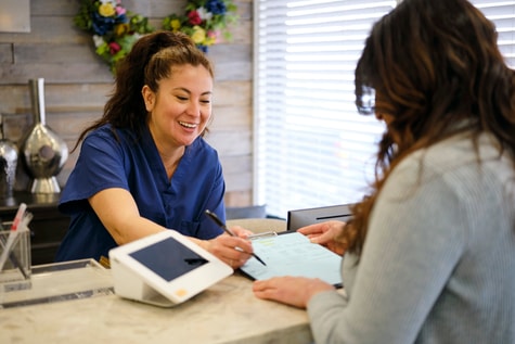 A smiling receptionist in scrubs helps a woman with paperwork at the front desk using a tablet running medbridge software.