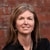 A woman with long hair in a black shirt smiles softly against a brick wall.