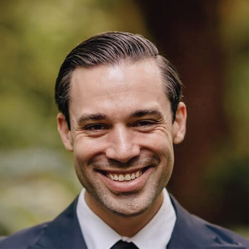 A smiling man in a suit and tie stands with a blurred natural backdrop.