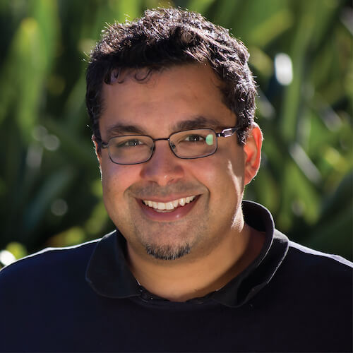 A person with glasses and short curly hair stands outside in front of green foliage, wearing a black shirt.