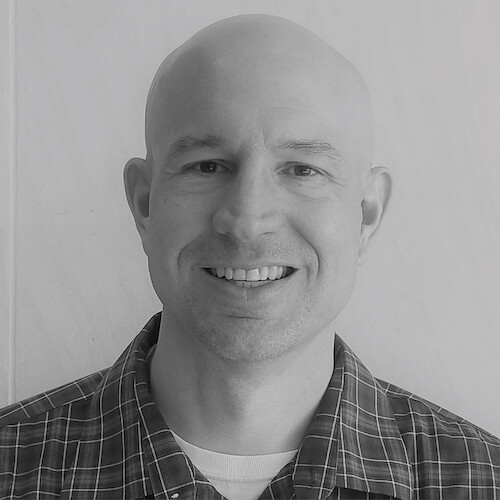 A smiling man with a shaved head in a plaid shirt, facing the camera in black and white.