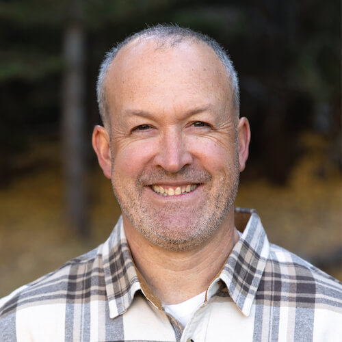 A man wearing a plaid shirt smiles at the camera against a blurred outdoor background for medbridge.