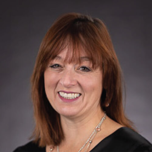 A woman with brown hair smiles in a black top, dark background.