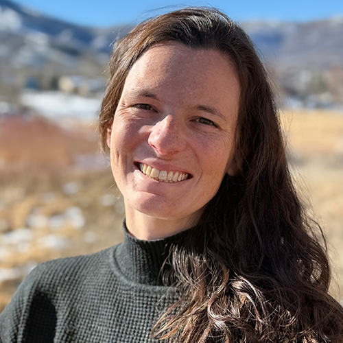 Smiling woman with long brown hair and black sweater outdoors, blurred mountains behind her, for medbridge hybrid care.