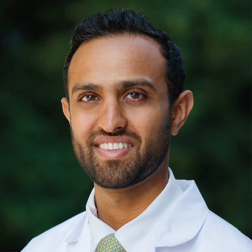 Man in a white coat smiling, with a dark green blurred background.