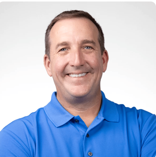 Smiling man with short brown hair in a blue polo shirt stands with arms crossed against a white background.