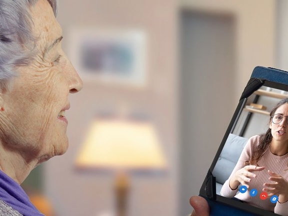 Elderly woman video chats on a tablet wearing a purple scarf in a cozy room using medbridge.