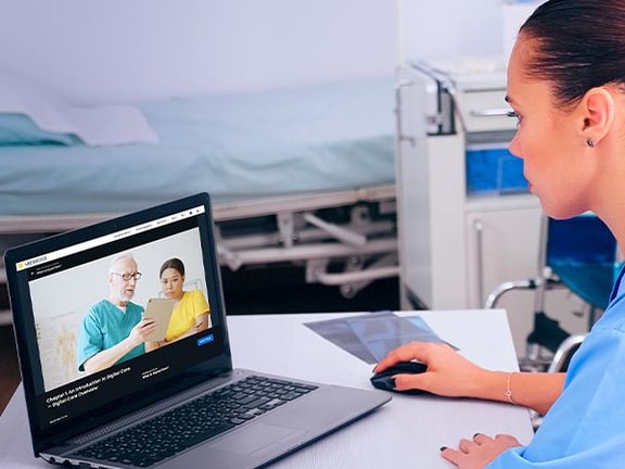 A healthcare professional in scrubs sits at a desk using medbridge on a laptop for a video call in a clinical setting.