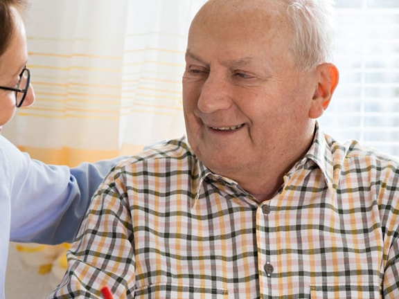 A woman in glasses converses with an older man in a plaid shirt near a window with white blinds.