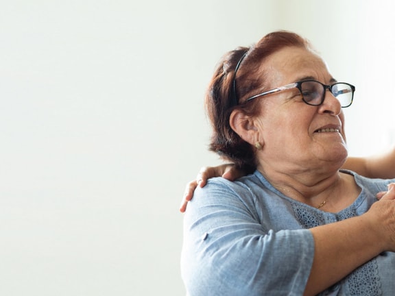 A caregiver in a white uniform converses with an older woman in a wheelchair against a plain background.