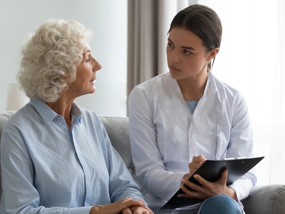 An elderly woman converses with a healthcare professional in a bright living room, with a wheelchair nearby.
