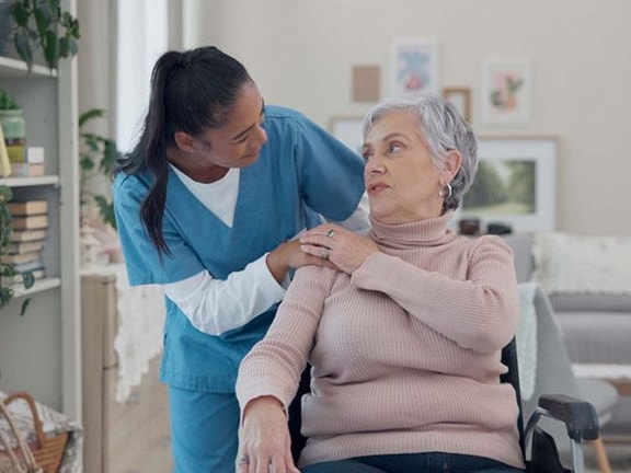 A caregiver in blue scrubs stands beside an elderly woman in a wheelchair, gently holding her shoulder.