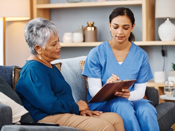 A nurse in blue scrubs talks with an older woman about health plans using medbridge software in a home-like room.