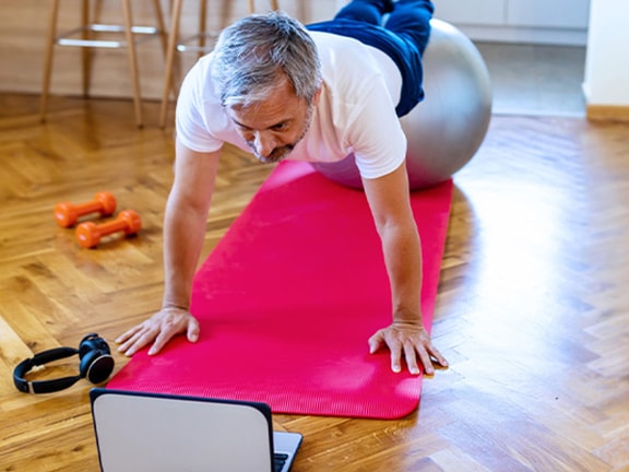 A person works out on a stability ball with a laptop, headphones, and dumbbells nearby on a red mat.