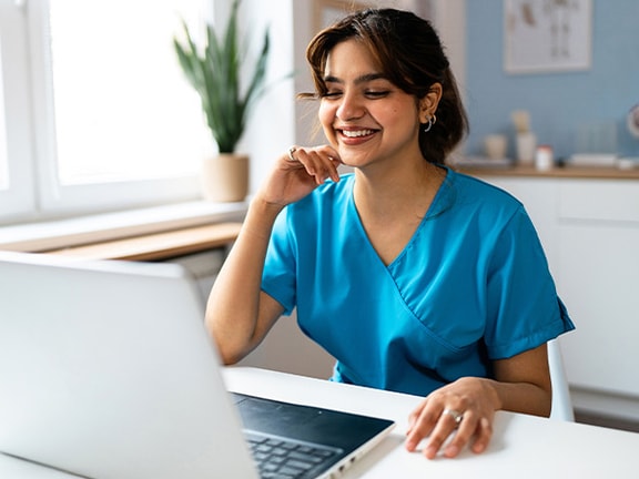 A person in blue medical scrubs smiles at a laptop, with a window and potted plant behind them.