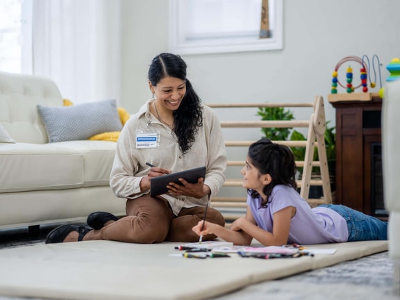Occupational therapist sitting on the floor with a young girl, using a tablet during a therapy session at home.