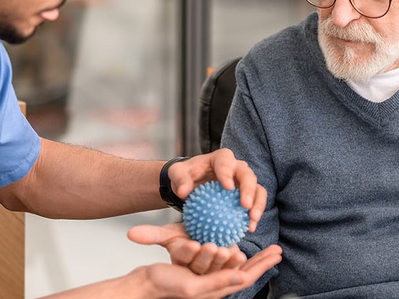 A Medbridge clinician in blue scrubs presents a textured object to an elderly man in a sweater.
