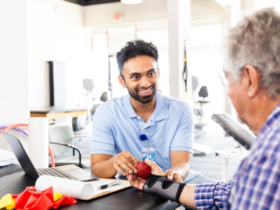 A healthcare provider helps an older adult do hand exercises with a red textured ball in a clinic.