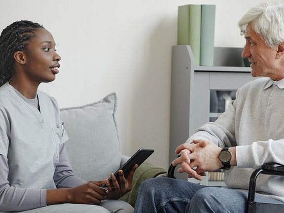 A Medbridge nurse with a tablet chats with a man in a wheelchair in a living room.