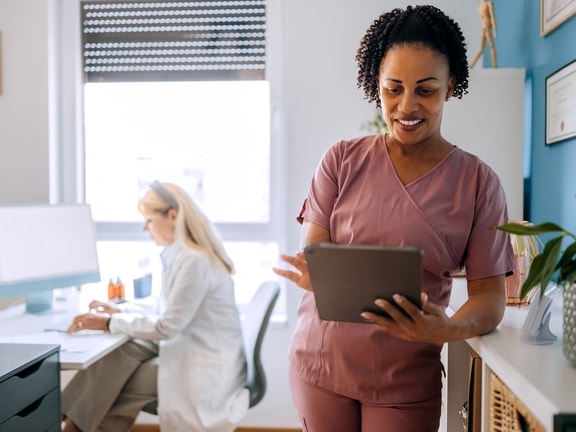 A healthcare worker in scrubs uses a tablet as another in a white coat works at a desk, showing medbridge digital health.