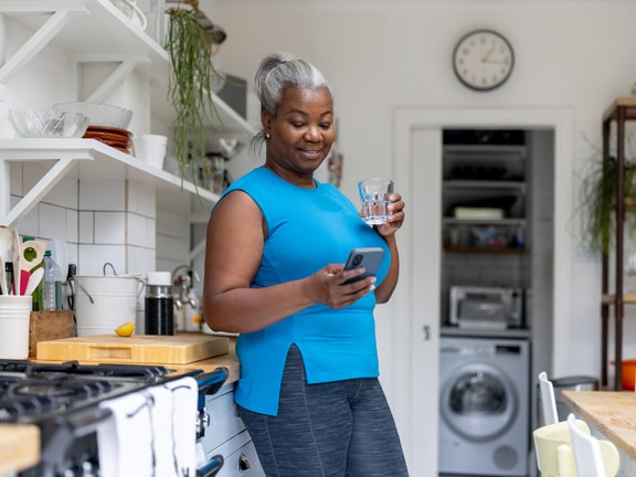 A woman in a blue top stands in a kitchen, holding a glass of water and looking at her phone, with kitchen appliances and plants behind her.