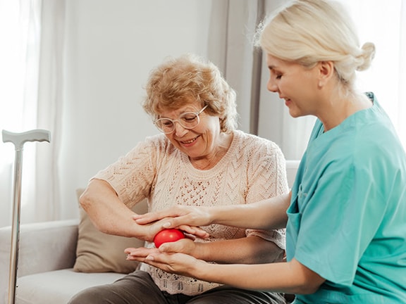 A nurse helps an elderly woman with hand exercises using a red ball on a couch.