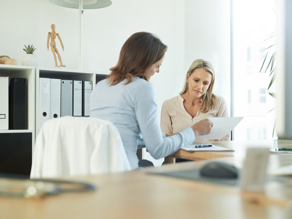 Two women review a document at a desk in an office with medbridge training materials in the background.